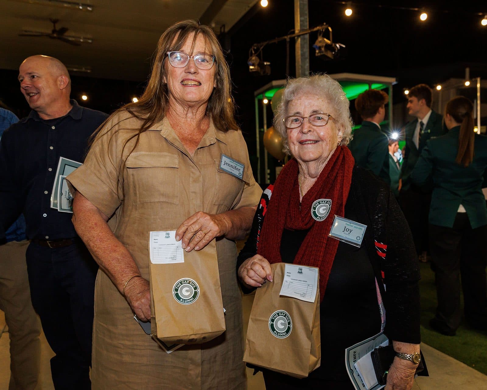 Alumni Launch: two guests holding gift bags smile for the camera, with students in school uniform behind them