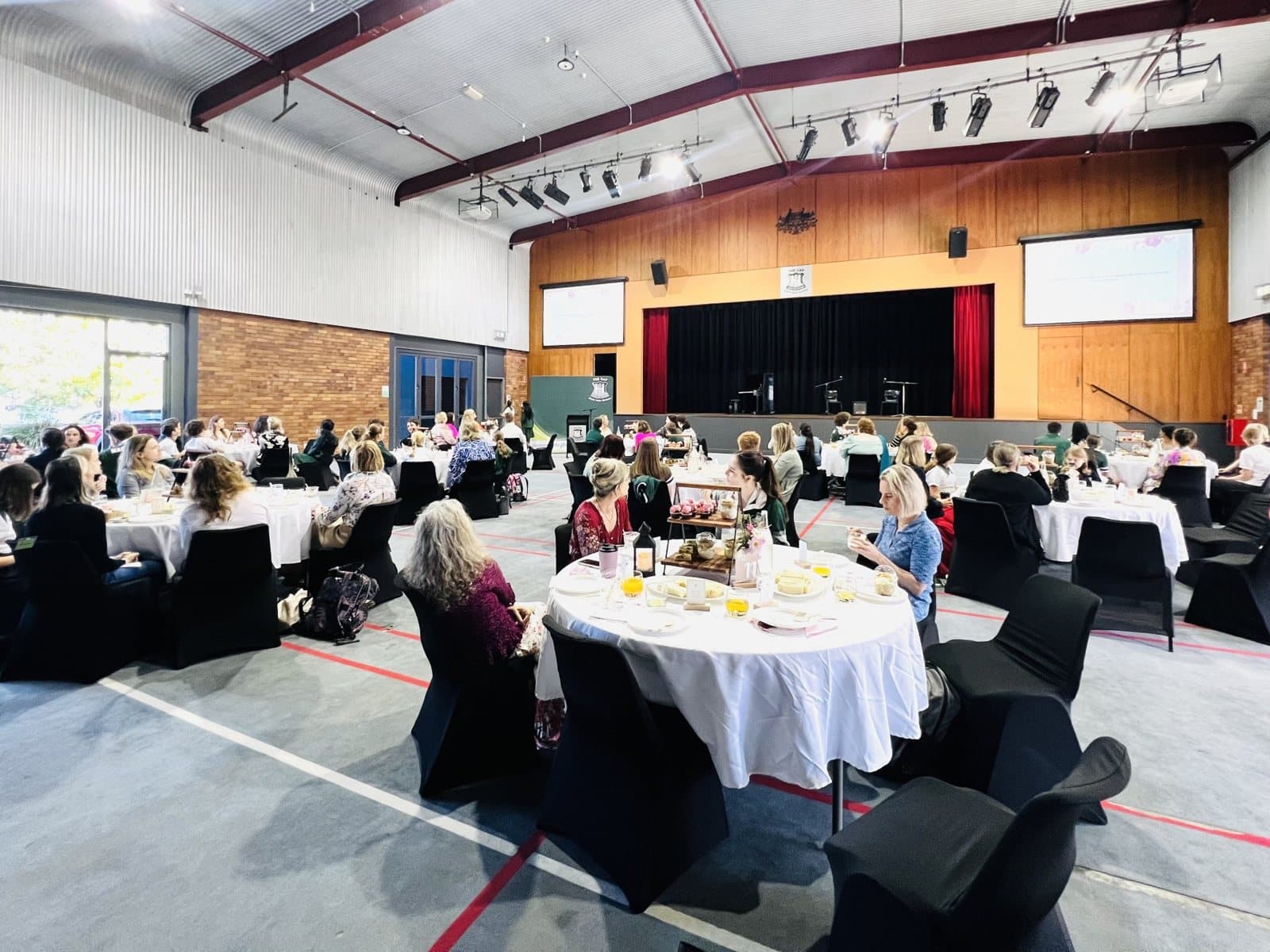 Mother's Day high tea: the school hall set with round tables in white cloths, guests seated and the stage at the far end