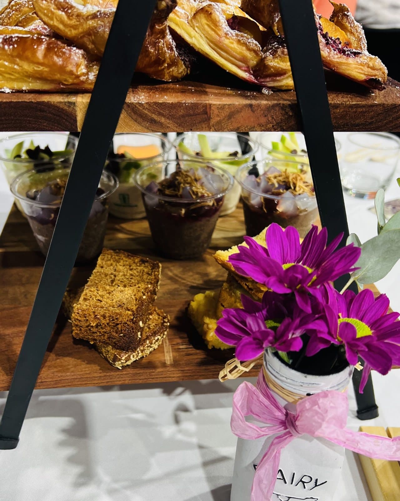 Mother's Day high tea: a tiered wooden stand of pastries and slices beside a jar of purple daisies tied with a pink ribbon